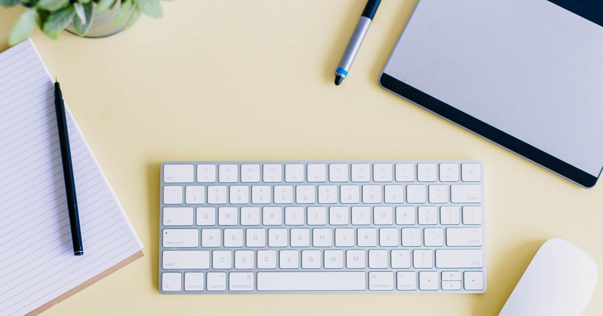 Top down image of a keyboard, mouse, notebook, pen, plant, on a yellow desktop surface.