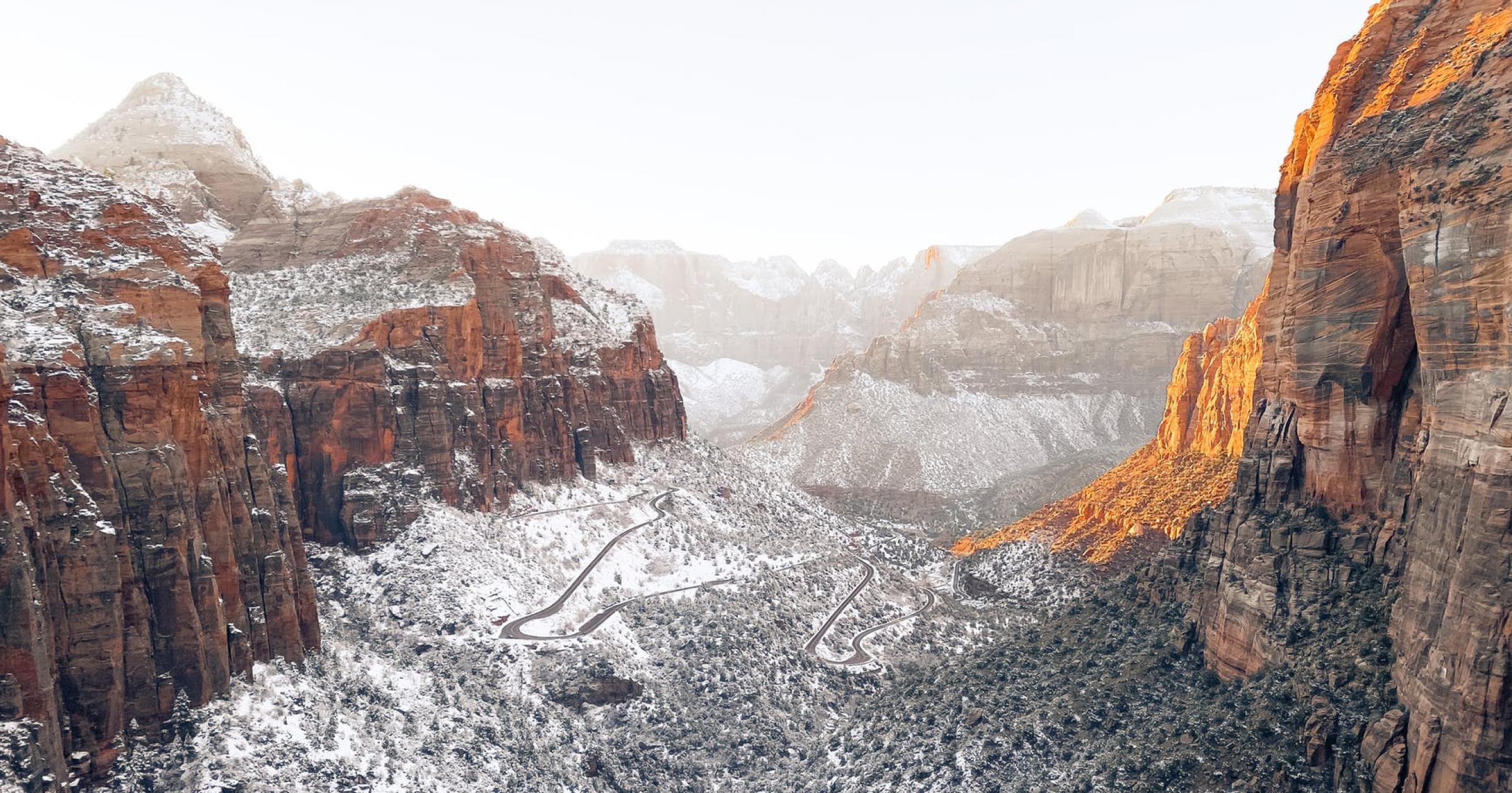 A wide landscape photo of a deep red rock canyon with a fresh dusting of snow and foggy clouds. A winding road on the canyon floor.